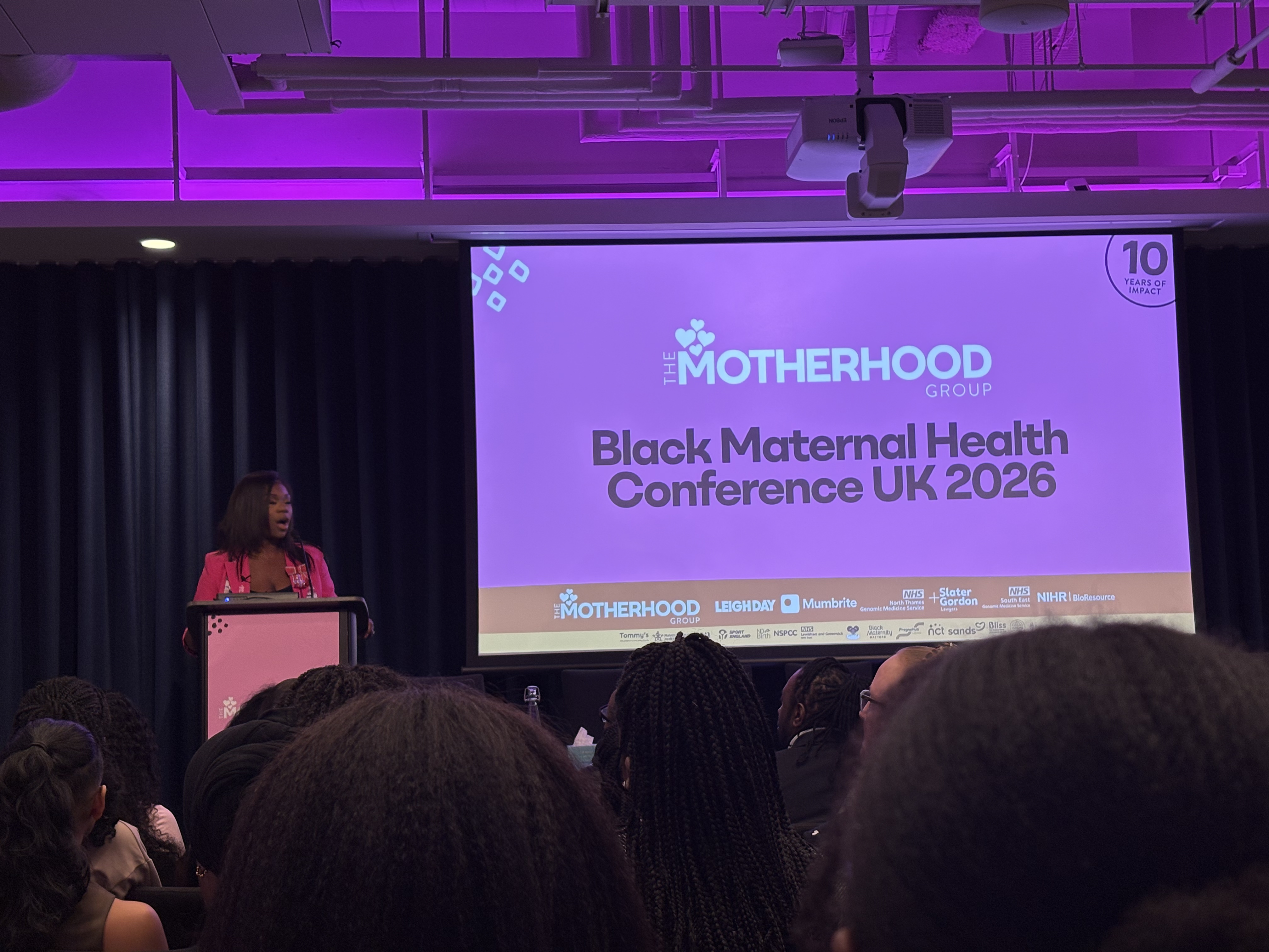 A Black woman wearing a pink suit stands at a podium. The presentaiton says "The Motherhood Group, Black Maternal Health Conference UK 2026"